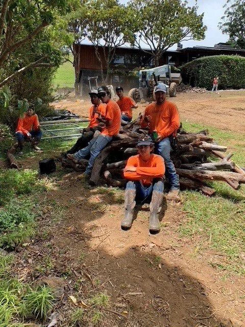 A group of people wearing orange shirts and hats sitting on a pile of wood in a rural setting.