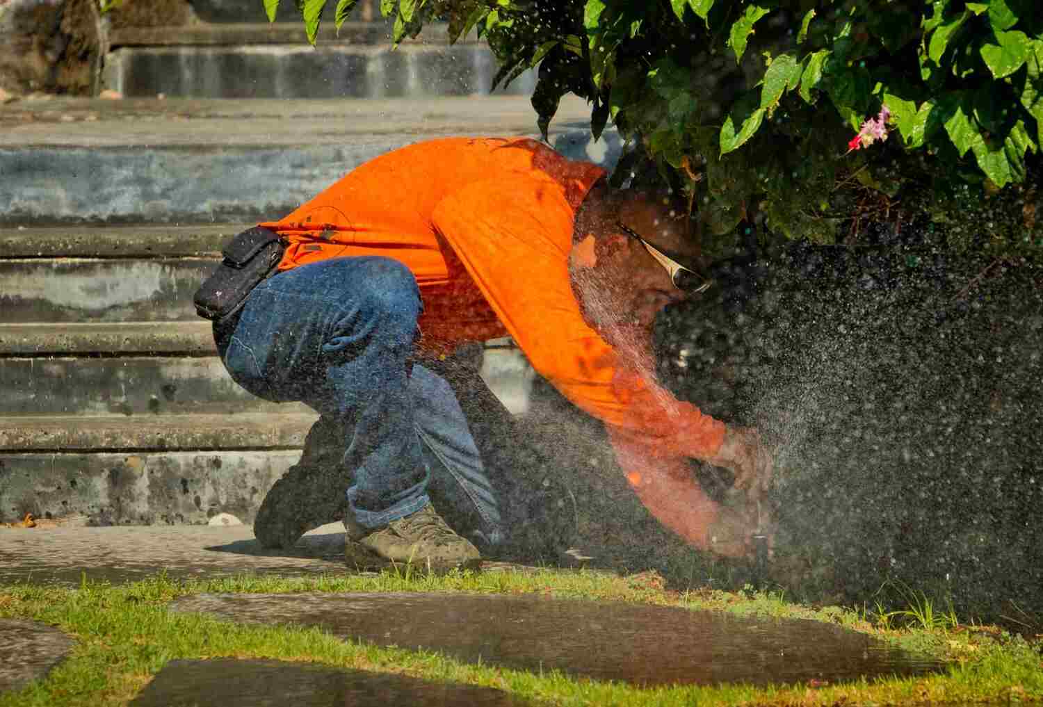 A man in an orange shirt kneeling on a lawn while adjusting a sprinkler system, with water spraying onto the grass.