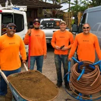 Four workers in bright orange shirts standing with a wheelbarrow and equipment, smiling at the camera. In the background, there are vehicles and coils of hose.