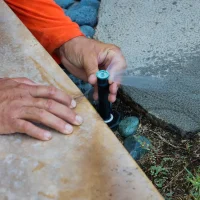 A person adjusting a sprinkler head on a lawn, with their hands visible on a stone surface.