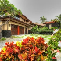 A view of a residential property featuring a two-story house surrounded by greenery, colorful flowers, and stone walls, set in a tropical environment.