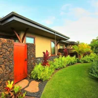A side view of a house surrounded by lush greenery, with a stone wall and a wooden door visible.
