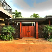A wooden gate surrounded by tropical plants and a stone wall, with a modern house visible on the left.